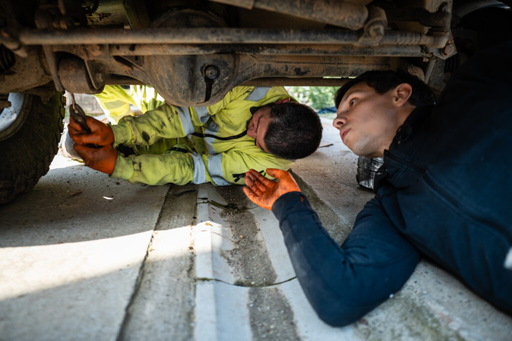 Two auto service mechanics lying on the ground, examining the undercarriage of a truck and discussing vehicle repair